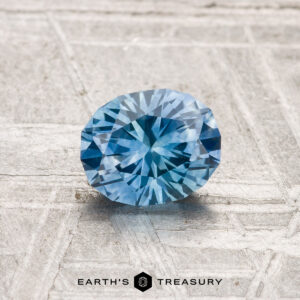A 1.01-carat heated Montana sapphire is showcased on a textured, light-colored surface with “EARTH’S TREASURY” displayed at the bottom.