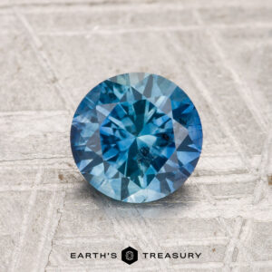 1.17-Carat Montana Sapphire (Heated) in round brilliant cut displayed on a textured light surface, featuring the text "EARTH'S TREASURY" at the bottom.