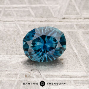 A faceted, oval-cut deep blue 1.01-carat heated Montana sapphire rests on a textured light gray background with "EARTH'S TREASURY" at the bottom.