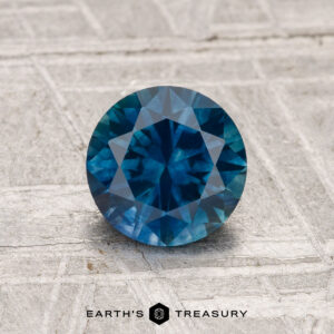 A 1.16-carat heated Montana sapphire, round and faceted, rests on a textured light gray surface with the "Earth's Treasury" logo at the bottom.