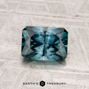 A rectangular, faceted teal-blue 1.35-carat Montana Sapphire (Heated) is displayed on a textured gray surface. The text "EARTH'S TREASURY" appears at the bottom of the image.