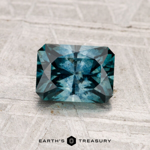 1.35-Carat Montana Sapphire (Heated) A rectangular, faceted teal-blue 1.35-carat Montana Sapphire (Heated) is displayed on a textured gray surface. The text "EARTH'S TREASURY" appears at the bottom of the image.