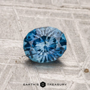 A 1.00-carat heated Montana sapphire is displayed on a textured gray surface with the "EARTH'S TREASURY" logo and text at the bottom.
