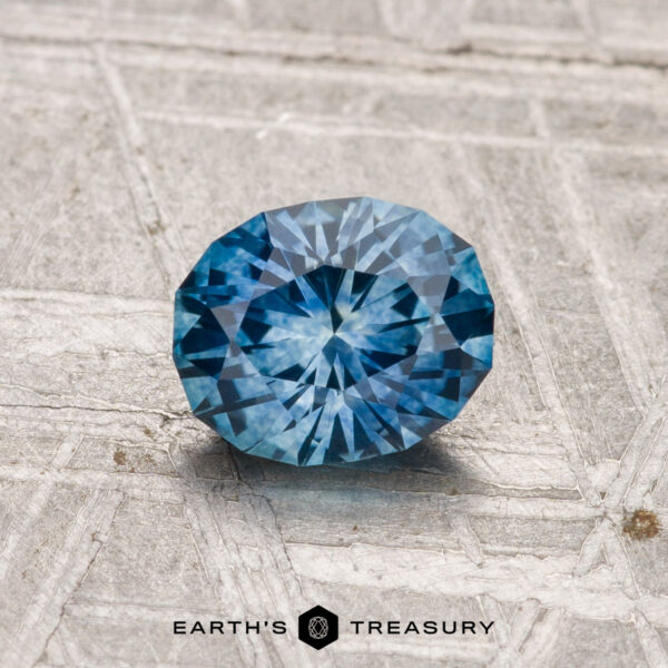 1.00-Carat Montana Sapphire (Heated) A 1.00-carat heated Montana sapphire is displayed on a textured gray surface with the "EARTH'S TREASURY" logo and text at the bottom.