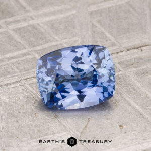 A 1.24-carat cushion-cut Ceylon sapphire rests on a textured gray surface with "Earth's Treasury" displayed at the bottom.