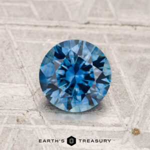 A 1.15-carat heated Montana Sapphire, round brilliant-cut and blue, is displayed on a metallic textured surface with the text "EARTH'S TREASURY" below.