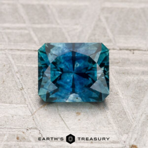 A 1.23-carat heated Montana sapphire with intricate faceting is displayed on a textured gray surface, featuring the Earth's Treasury logo at the bottom.