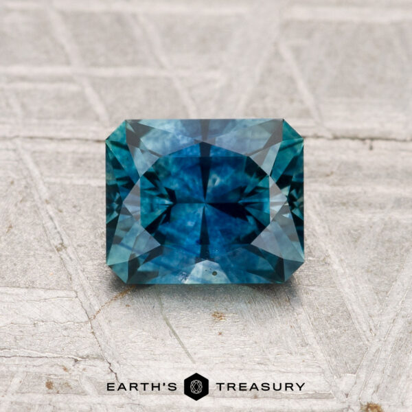 1.23-Carat Montana Sapphire (Heated) A 1.23-carat heated Montana sapphire with intricate faceting is displayed on a textured gray surface, featuring the Earth's Treasury logo at the bottom.