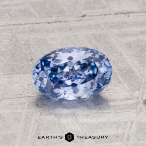 A 1.29-carat Ceylon sapphire with an oval cut and light blue hue rests on a textured gray surface, featuring "Earth's Treasury" branding at the bottom of the image.