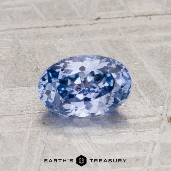 1.29-Carat Ceylon Sapphire A 1.29-carat Ceylon sapphire with an oval cut and light blue hue rests on a textured gray surface, featuring "Earth's Treasury" branding at the bottom of the image.