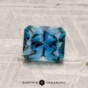 A 1.16-carat rectangular step-cut heated Montana sapphire is displayed on a textured gray surface with "EARTH'S TREASURY" text at the bottom.