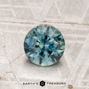 A round, faceted blue-green 1.05-carat Montana Sapphire (Heated) rests on a textured surface, with the "EARTH'S TREASURY" text and a hexagon logo beneath it.