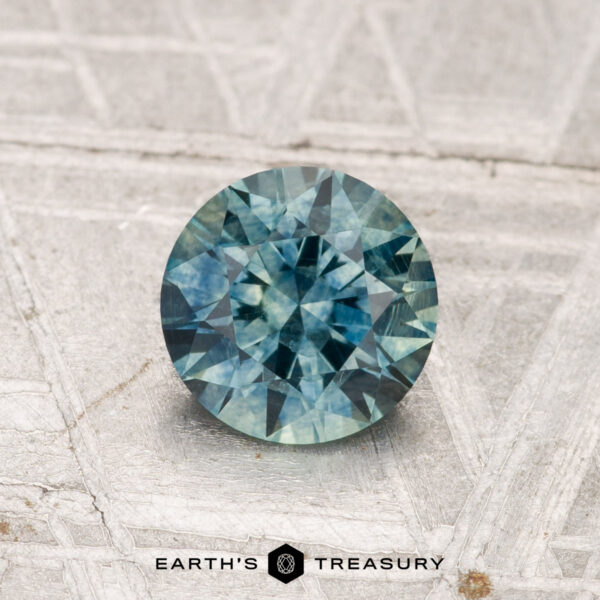 1.05-Carat Montana Sapphire (Heated) A round, faceted blue-green 1.05-carat Montana Sapphire (Heated) rests on a textured surface, with the "EARTH'S TREASURY" text and a hexagon logo beneath it.
