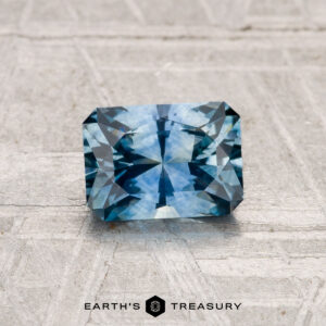 A rectangular, faceted 1.13-carat heated Montana sapphire is showcased on a textured light gray surface with "EARTH'S TREASURY" written beneath it.