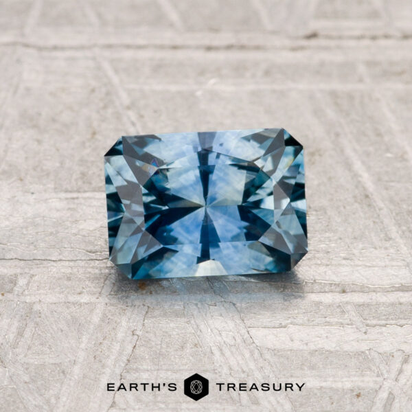 1.13-Carat Montana Sapphire (Heated) A rectangular, faceted 1.13-carat heated Montana sapphire is showcased on a textured light gray surface with "EARTH'S TREASURY" written beneath it.