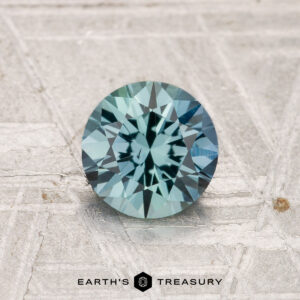 A 1.05-carat heated Montana sapphire with a round brilliant cut and blue-green color is displayed on a textured gray surface, with "Earth's Treasury" shown at the bottom.