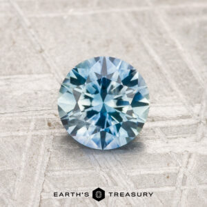 A 0.99-carat, round brilliant-cut heated Montana sapphire is displayed on a textured light gray surface, with the "EARTH'S TREASURY" logo and text at the bottom of the image.