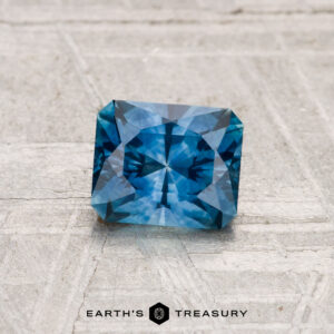 A 1.05-carat heated Montana sapphire, expertly square-cut with intricate facets, is shown on a textured light gray surface. "EARTH'S TREASURY" is displayed at the bottom.