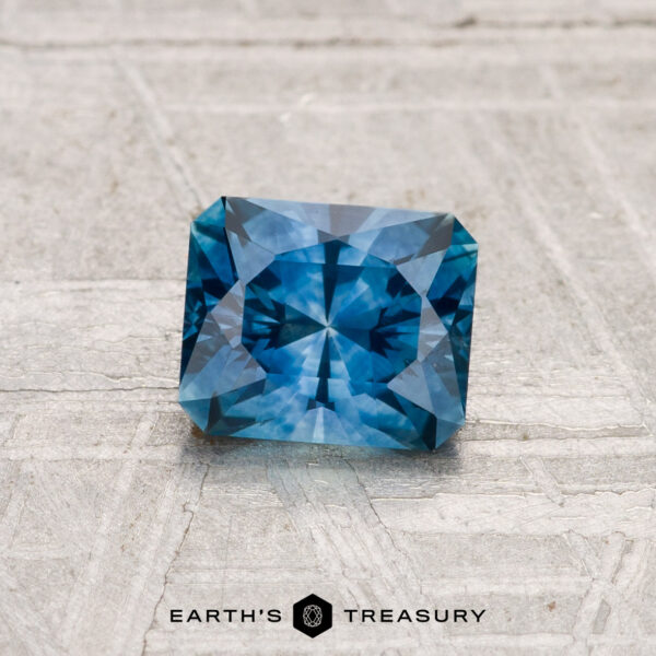 1.05-Carat Montana Sapphire (Heated) A 1.05-carat heated Montana sapphire, expertly square-cut with intricate facets, is shown on a textured light gray surface. "EARTH'S TREASURY" is displayed at the bottom.