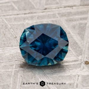 A 1.96-carat faceted Montana sapphire (heated) in deep blue rests on a textured grey surface, with "EARTH'S TREASURY" displayed at the bottom.