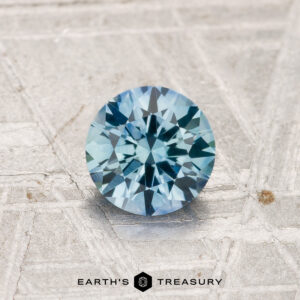 A 0.94-carat heated Montana sapphire, cut in a round brilliant shape, is shown on a textured surface with "EARTH'S TREASURY" at the bottom.
