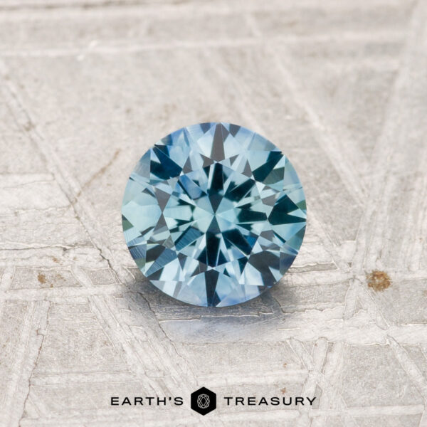 0.94-Carat Montana Sapphire (Heated) A 0.94-carat heated Montana sapphire, cut in a round brilliant shape, is shown on a textured surface with "EARTH'S TREASURY" at the bottom.