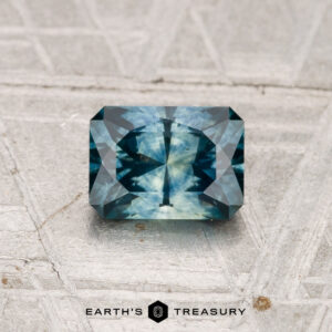 A 1.02-carat heated Montana sapphire, rectangular and blue-green with intricate facets, is displayed on a textured grey surface, with "Earth's Treasury" branding below.