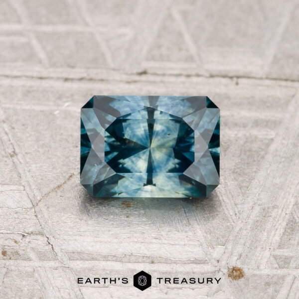 1.02-Carat Montana Sapphire (Heated) A 1.02-carat heated Montana sapphire, rectangular and blue-green with intricate facets, is displayed on a textured grey surface, with "Earth's Treasury" branding below.