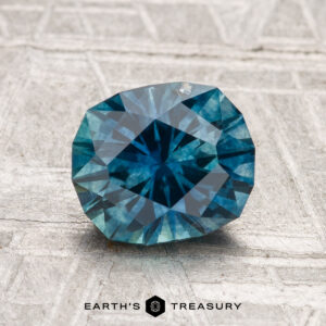 A 1.84-carat heated Montana sapphire rests on a textured gray surface, with "EARTH'S TREASURY" visible below.