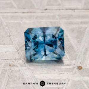A 0.99-carat heated Montana Sapphire, square-cut and blue, sits on a textured, light-colored surface with the Earth’s Treasury logo beneath it.