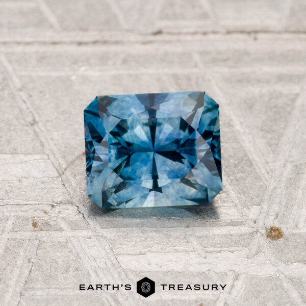 0.99-Carat Montana Sapphire (Heated) A 0.99-carat heated Montana Sapphire, square-cut and blue, sits on a textured, light-colored surface with the Earth’s Treasury logo beneath it.