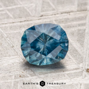 A 1.61-carat heated Montana sapphire rests on a textured metallic surface, with “EARTH'S TREASURY” and a geometric logo displayed below.