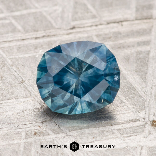 1.61-Carat Montana Sapphire (Heated) A 1.61-carat heated Montana sapphire rests on a textured metallic surface, with “EARTH'S TREASURY” and a geometric logo displayed below.