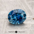 A faceted blue 1.43-carat Montana sapphire (heated) rests on a textured gray surface; "EARTH'S TREASURY" logo and text appear at the bottom.