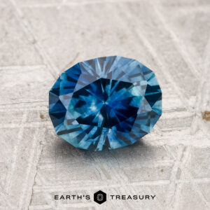 A faceted blue 1.43-carat Montana sapphire (heated) rests on a textured gray surface; "EARTH'S TREASURY" logo and text appear at the bottom.