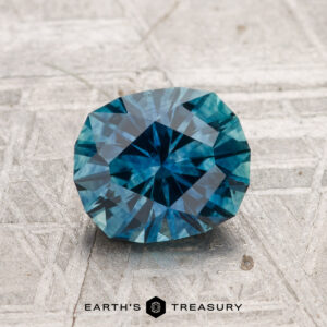 A 1.61-carat heated Montana sapphire with geometric facets is displayed on a textured gray surface. "EARTH'S TREASURY" appears at the bottom.