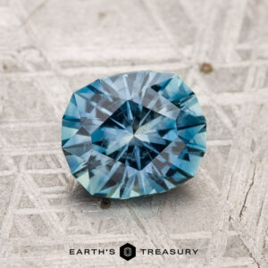 A 1.57-carat heated Montana sapphire is shown on a patterned metallic surface with "Earth's Treasury" visible at the bottom.
