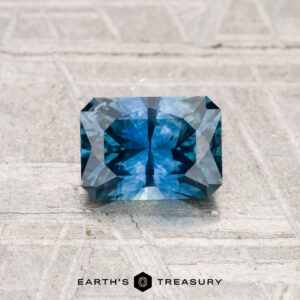 A 0.97-carat heated Montana sapphire with intricate faceting is displayed on a textured light gray surface. "EARTH'S TREASURY" appears at the bottom.