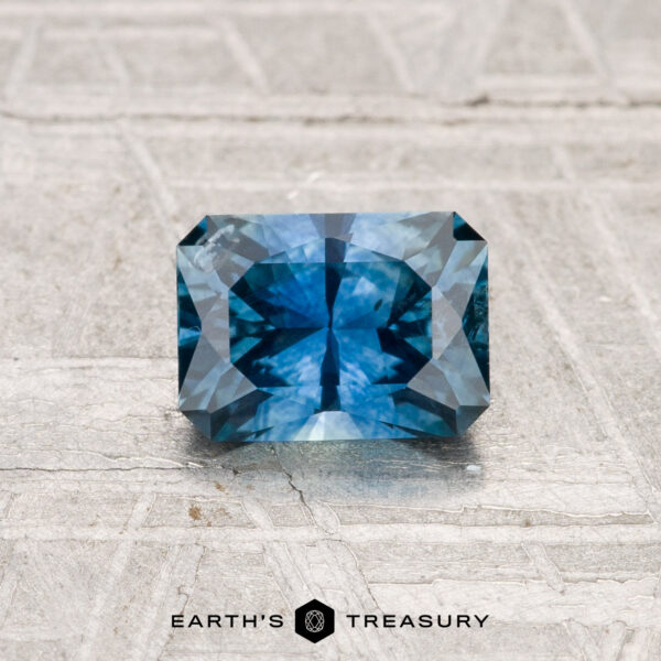 0.97-Carat Montana Sapphire (Heated) A 0.97-carat heated Montana sapphire with intricate faceting is displayed on a textured light gray surface. "EARTH'S TREASURY" appears at the bottom.