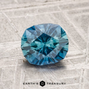 A 1.52-carat heated Montana sapphire with a blue octagonal facet rests on a textured gray surface; the "Earth's Treasury" logo appears at the bottom.