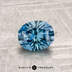 1.24-Carat Montana Sapphire (Heated) with a faceted blue starburst pattern, displayed on a textured light gray surface, features the "Earth's Treasury" logo at the bottom.