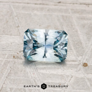 A rectangular, faceted light blue 0.93-carat Montana sapphire (heated) is displayed on a textured gray surface with the text "EARTH'S TREASURY" at the bottom.