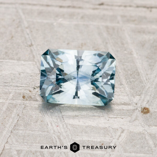 0.93-Carat Montana Sapphire (Heated) A rectangular, faceted light blue 0.93-carat Montana sapphire (heated) is displayed on a textured gray surface with the text "EARTH'S TREASURY" at the bottom.