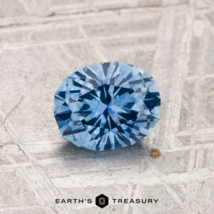 A 1.21-carat heated Montana sapphire in a round brilliant cut is displayed on a textured gray surface with "EARTH'S TREASURY" at the bottom.