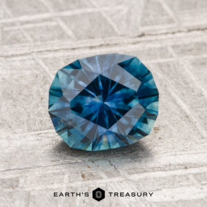 A 1.45-carat heated Montana Sapphire is displayed on a textured gray surface, with "EARTH'S TREASURY" visible at the bottom.