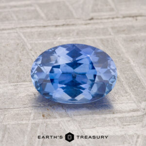 A 1.54-carat Ceylon sapphire, oval-cut and blue, is shown on a textured gray surface with "EARTH'S TREASURY" written below.