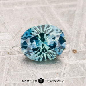 A round, faceted blue-green 1.17-carat Montana Sapphire (Heated) rests on a textured, light-colored surface, with "EARTH'S TREASURY" displayed at the bottom.