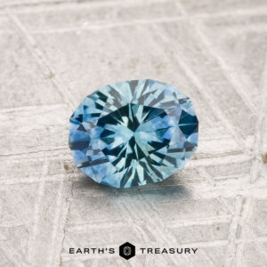 A 1.11-carat heated Montana sapphire, round brilliant-cut, is displayed on a textured gray surface with "EARTH'S TREASURY" at the bottom.