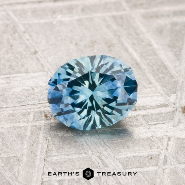 1.11-Carat Montana Sapphire (Heated) A 1.11-carat heated Montana sapphire, round brilliant-cut, is displayed on a textured gray surface with "EARTH'S TREASURY" at the bottom.