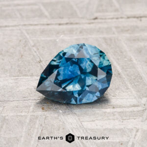 A 1.04-carat heated Montana sapphire, faceted in a pear shape, rests on a light gray textured surface. The "EARTH'S TREASURY" logo is displayed at the bottom of the image.